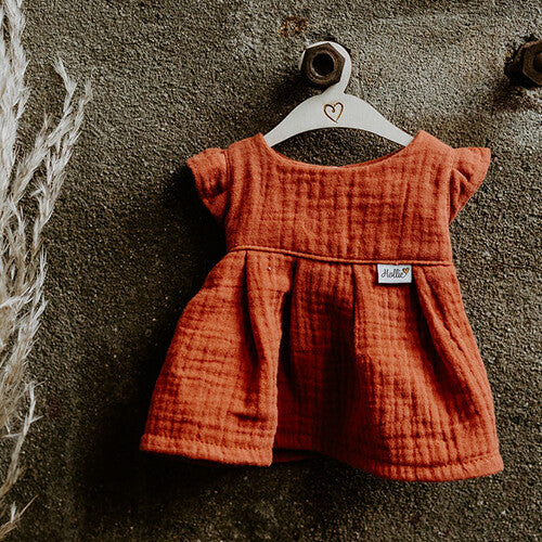 Hollie poppenkleding jurkje in terra katoen met textuur, hangend aan een houten hanger tegen een donkere muur.