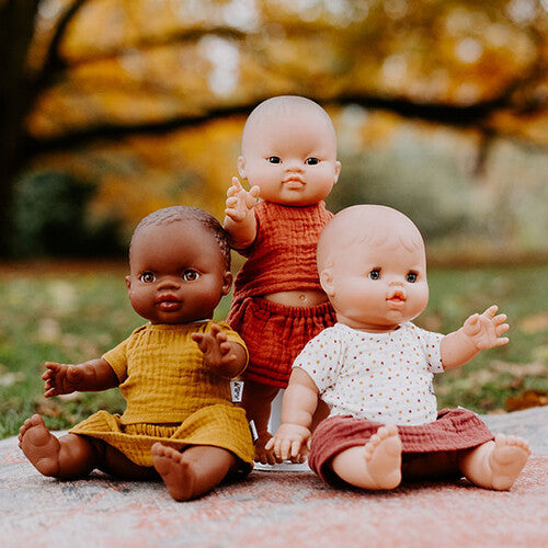 Drie babypoppen met verschillende huidskleuren dragen okergele en roodbruine poppenkleding buiten op een kleed.