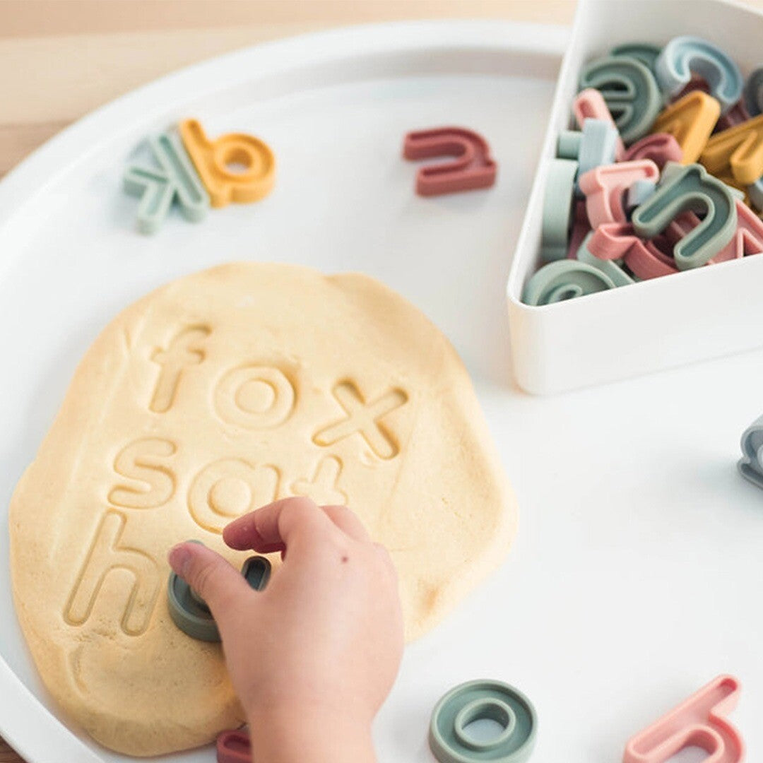 Een kind drukt een inspire my play siliconen letter in klei, met andere letters en een bakje met letters op een witte ondergrond.