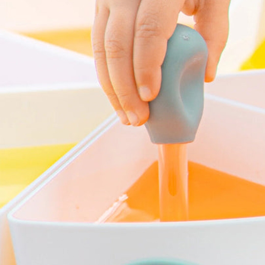Een kinderhand gebruikt een grijze pipet om oranje vloeistof uit een witte bak te zuigen.