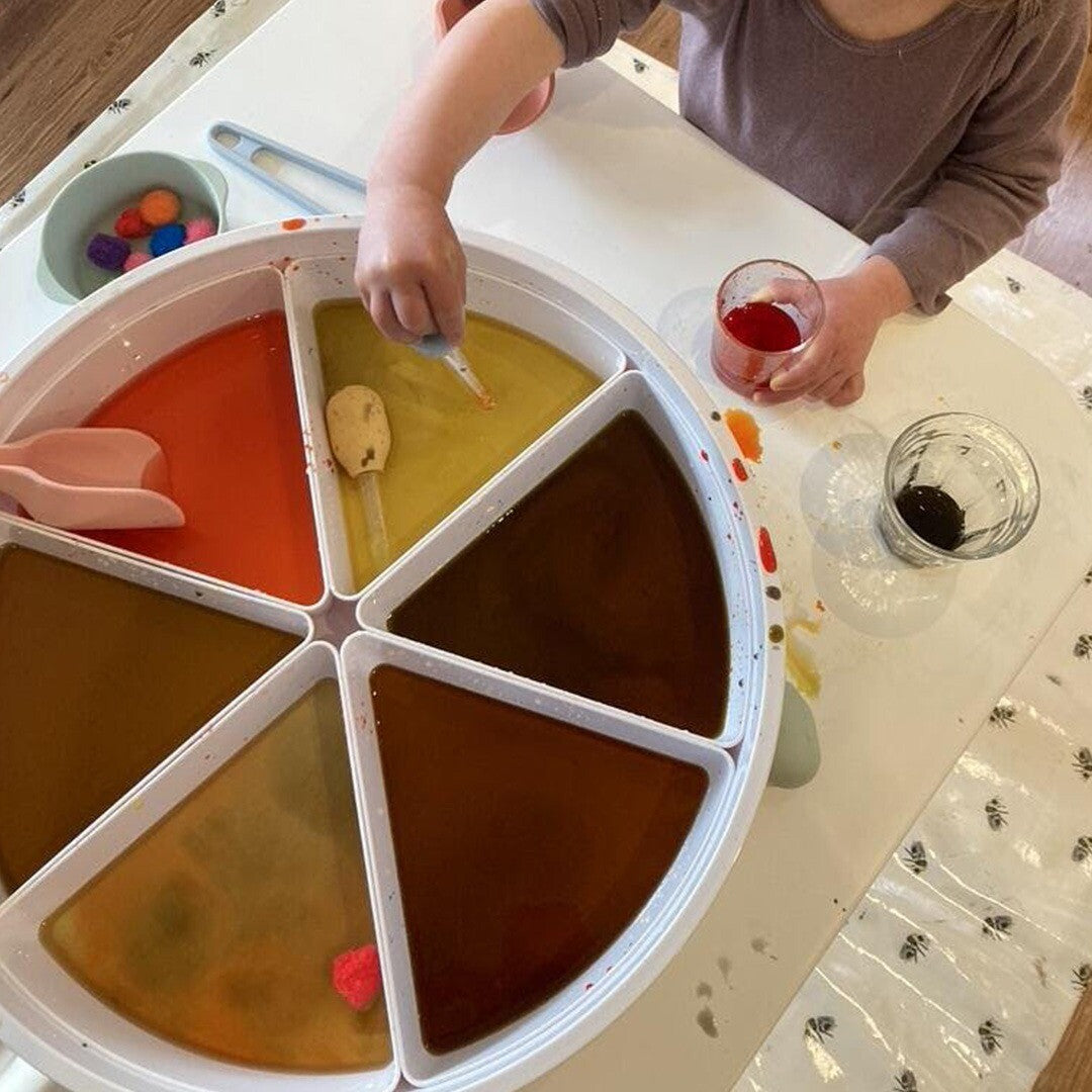 Een kind speelt met een pipet in een grote ronde bak met verschillende gekleurde vloeistoffen.