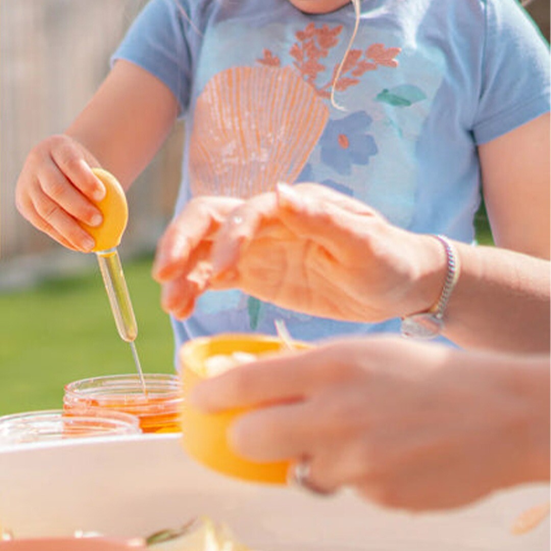 Een kinderhand gebruikt een gele pipet om vloeistof in een klein bakje te druppelen.