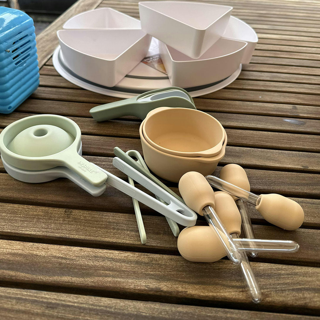 Een collectie inspire my play sensorisch speelgoed, waaronder beige pipetten, bakjes en tangen, ligt op een houten tafel.