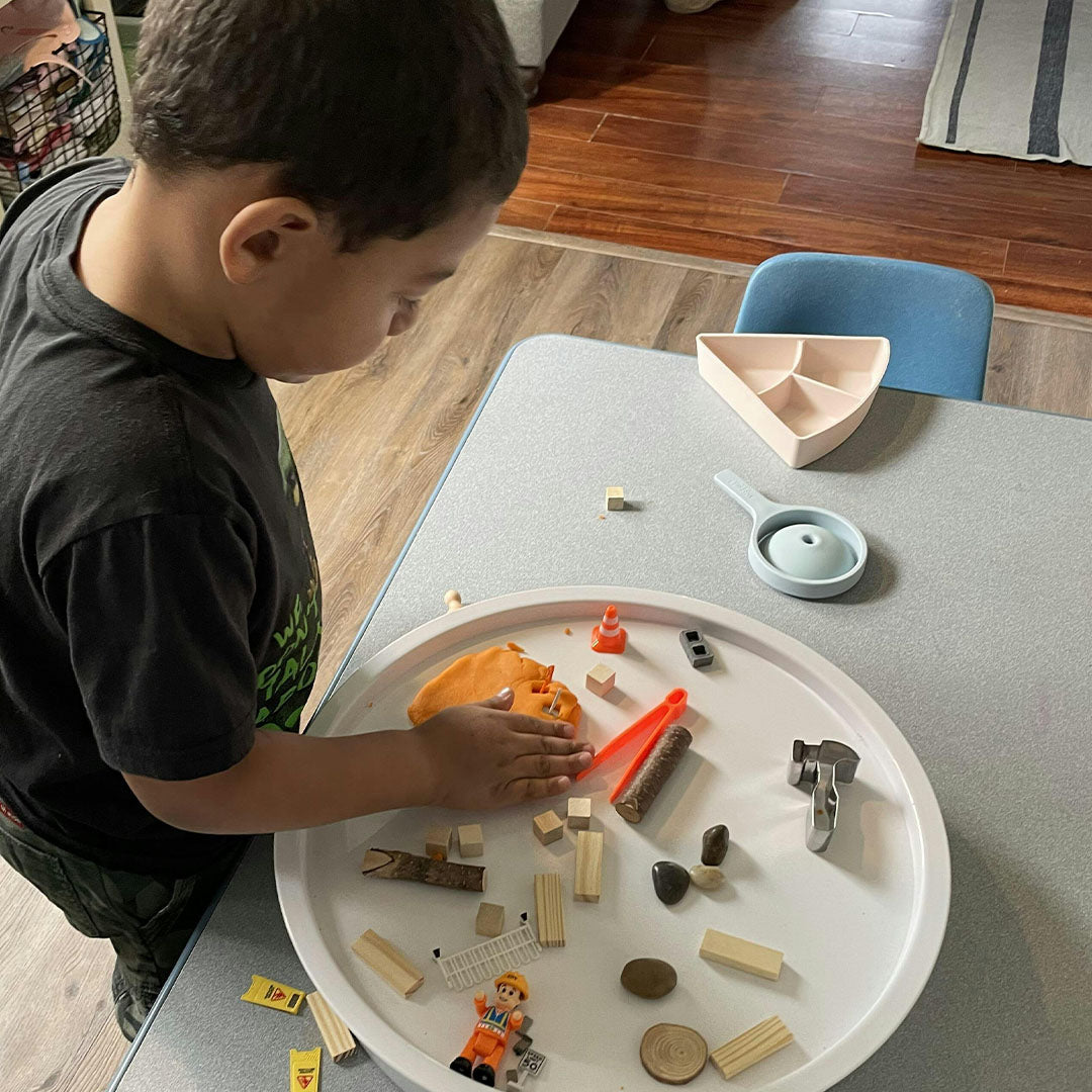 Een kind speelt aan tafel met een grote ronde speelbak gevuld met sensorisch speelgoed en een beige playtray insert op de achtergrond.
