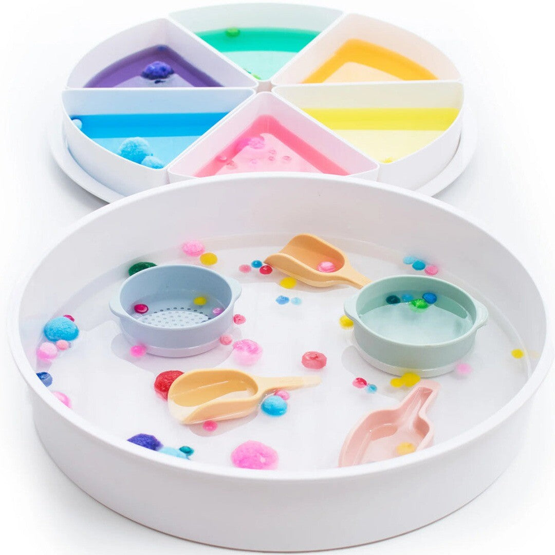 inspire my play speelbak gevuld met water, kleurrijke pompons en schepjes, met een gesegmenteerde tray met gekleurde vloeistoffen erachter.
