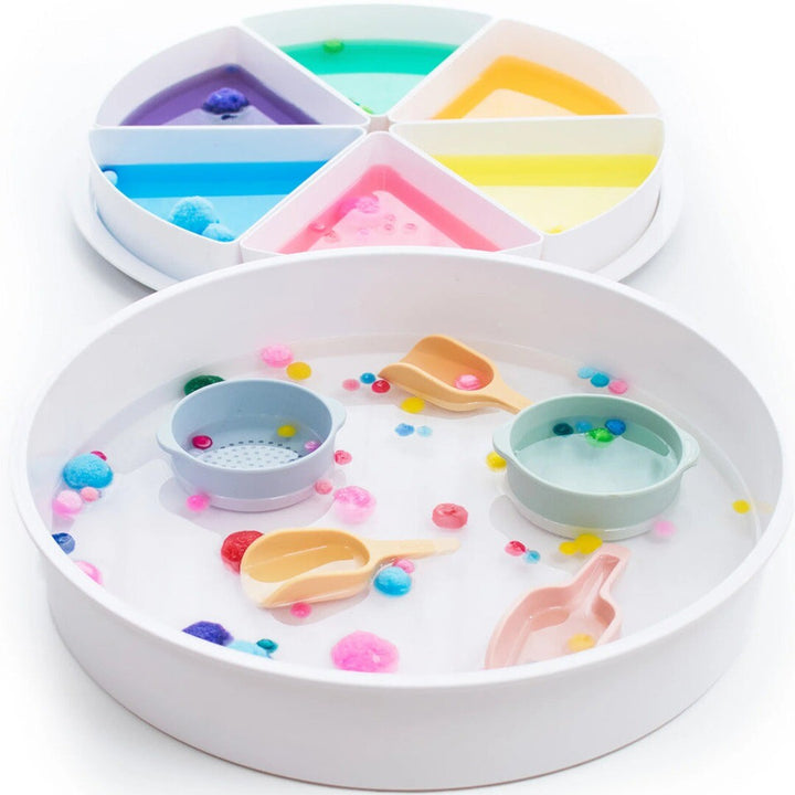 inspire my play speelbak gevuld met water, kleurrijke pompons en schepjes, met een gesegmenteerde tray met gekleurde vloeistoffen erachter.