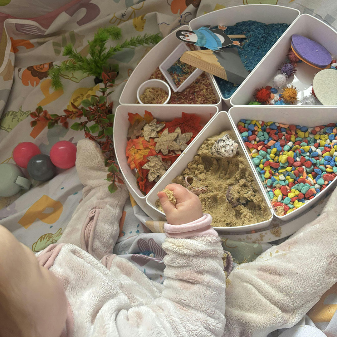Een babyhandje reikt naar zand en natuurlijke materialen in een inspire my play gesegmenteerde speelbak.