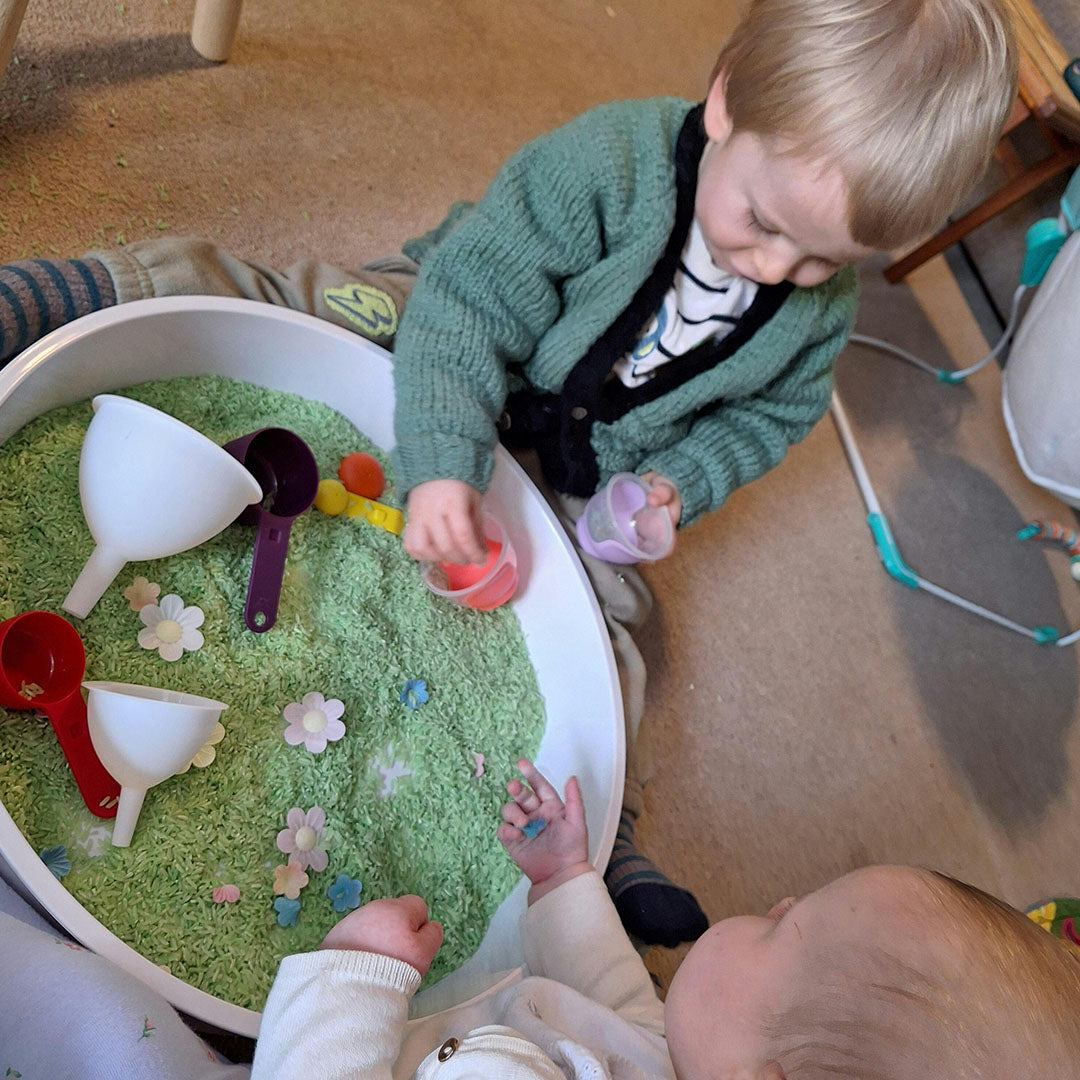 Twee kinderen spelen met een inspire my play speelbak gevuld met groene rijst, bloemetjes, trechters en schepjes.
