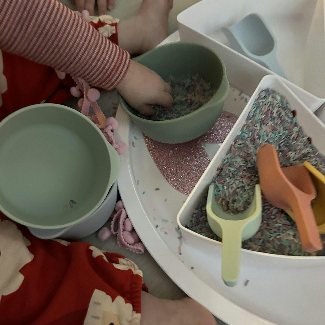 Een kind speelt met inspire my play sensorische schepjes en bakjes gevuld met gekleurde rijst.