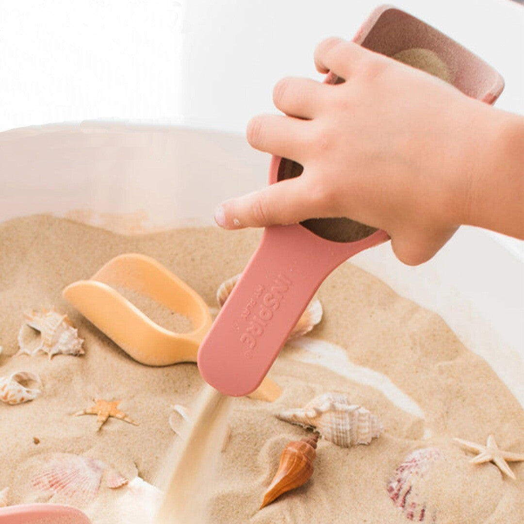 Een kinderhand giet zand uit een roze inspire my play sensorisch schepje in een witte bak met zeeschelpen.