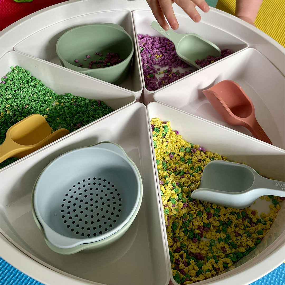 Een kinderhand reikt in een ronde sorteerbak met kleurrijke sensorische materialen en inspire my play siliconen sorteerbakjes en schepjes.