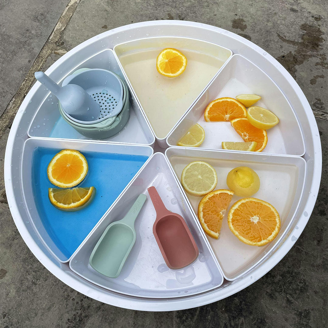 Een grote ronde witte sorteerbak met zes vakken, gevuld met water, citroen- en sinaasappelschijfjes, en inspire my play sorteerbakjes en schepjes.