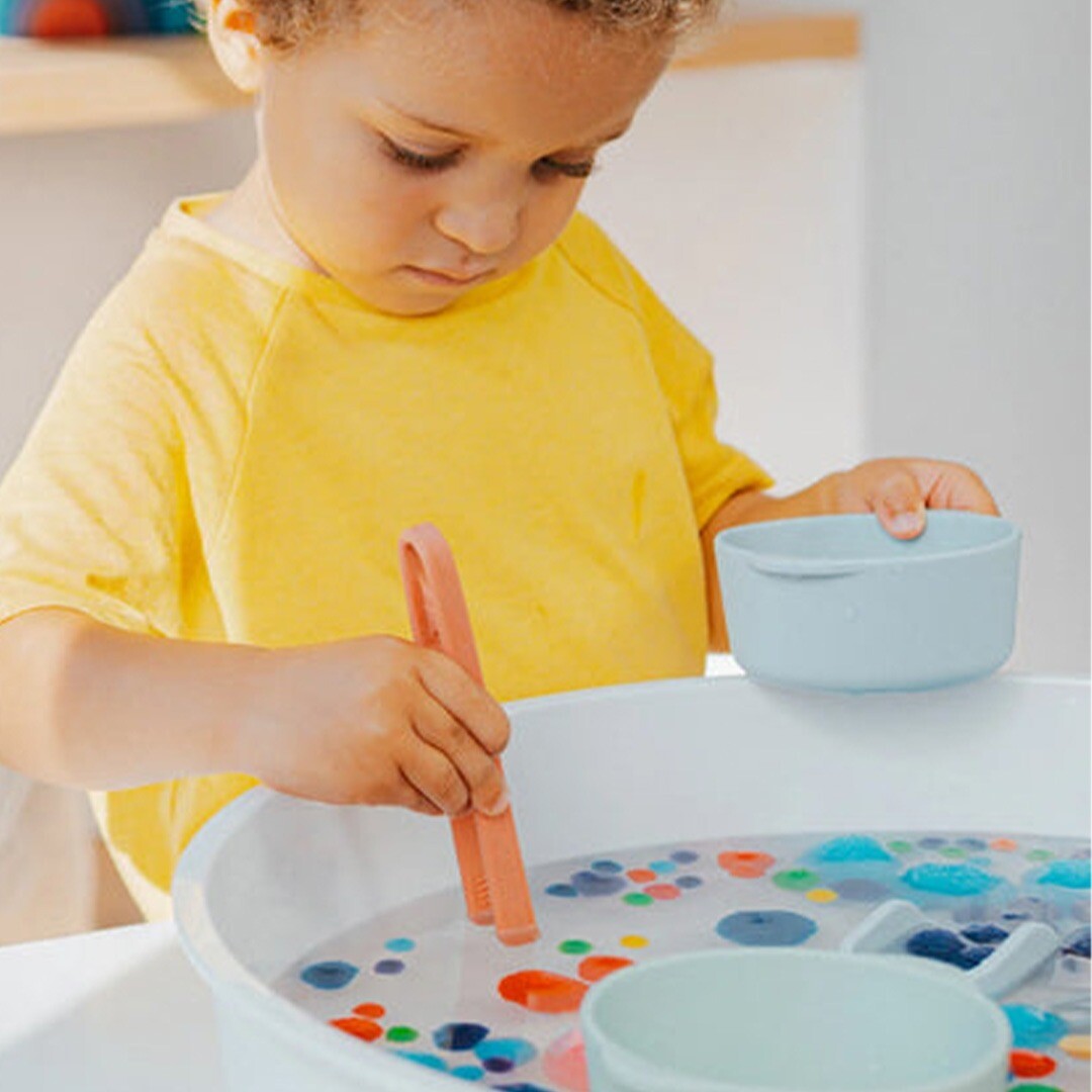 Een kind gebruikt een koraalkleurige inspire my play tang om drijvende voorwerpen uit een bak met water te pakken.