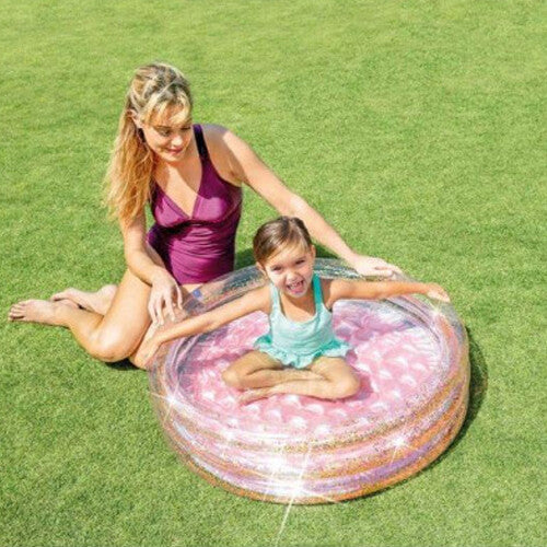 Een lachend meisje zit in het Intex glitter peuterzwembad op gras, met een volwassene ernaast.