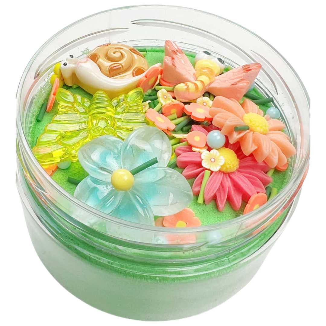 Invitation to imagine magic sand surprise pot met tuinthema, met een slak, vlinders en bloemen in groen zand, schuin van bovenaf gezien.