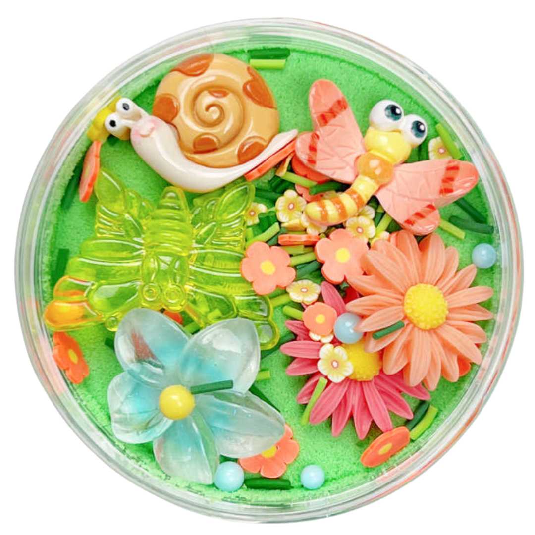 Invitation to imagine magic sand surprise pot met tuinthema, met een slak, vlinders en bloemen in groen zand, van bovenaf gezien.