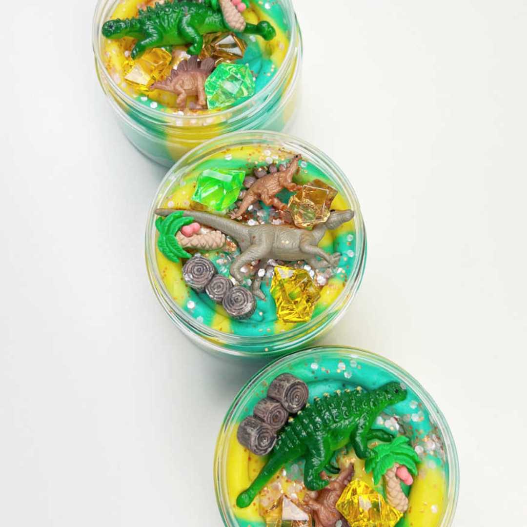 Drie invitation to imagine speelklei surprise potten met dinosaurusfiguren, palmboompjes en glitters in gele en blauwgroene klei.