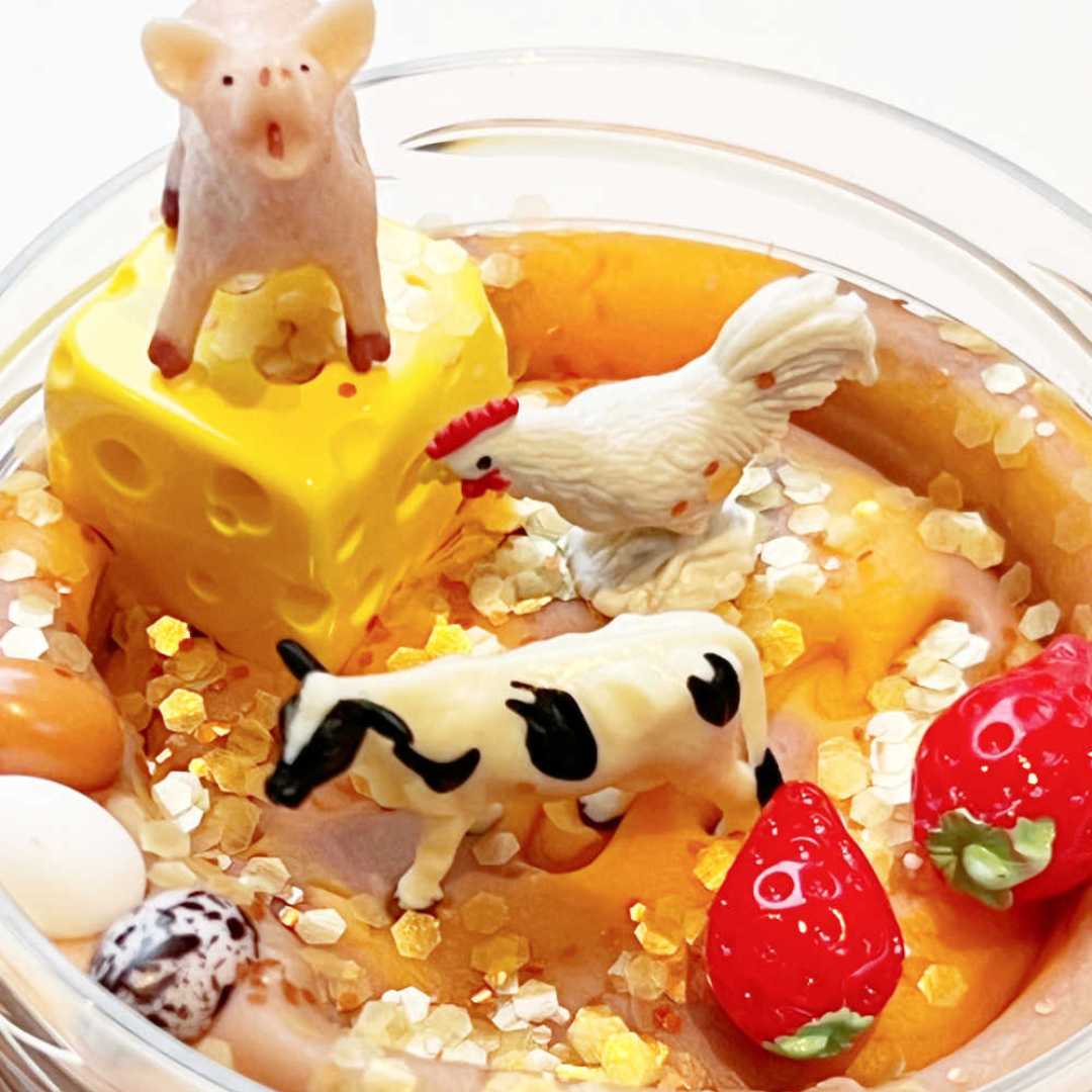 Detailfoto van de invitation to imagine speelklei pot met boerderijfiguren, kaas, aardbeien en snoepjes in gele en bruine klei.
