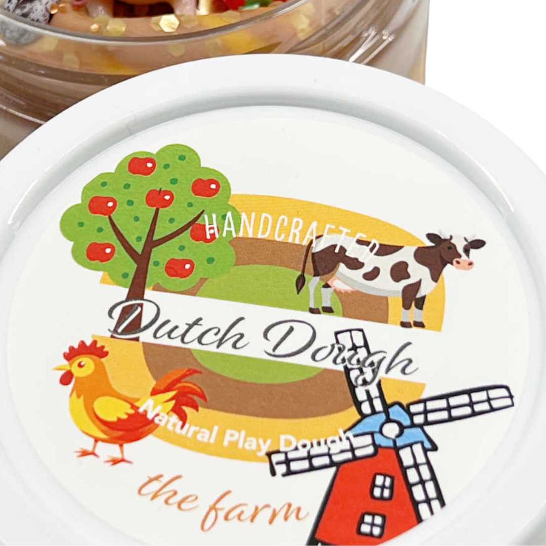 De witte deksel van de invitation to imagine speelklei pot met een boerderij-illustratie van een koe, haan, appelboom en molen.