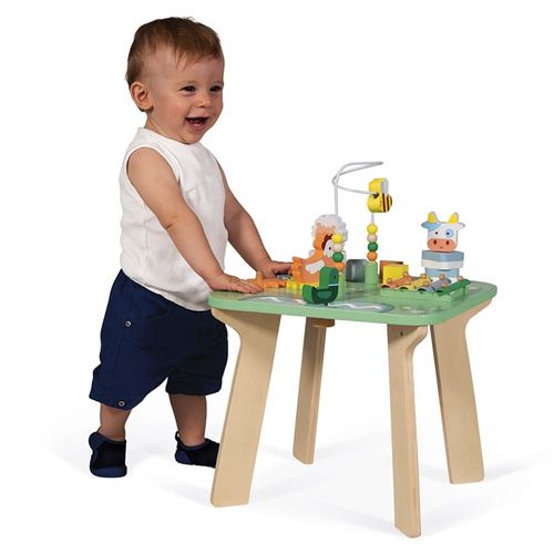 Een lachende baby speelt staand met de Janod houten activiteitenspeeltafel 'De Weide'.