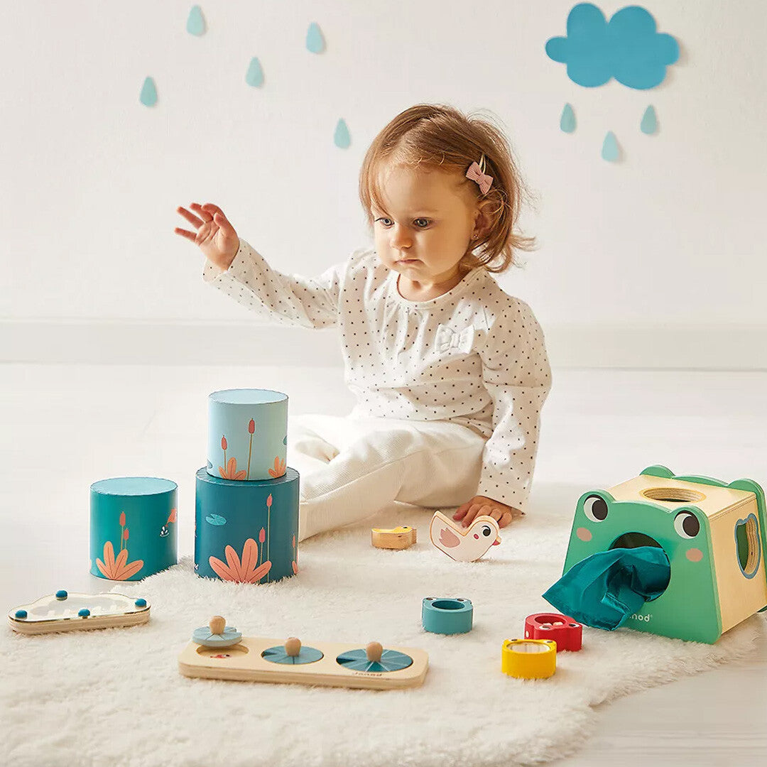 Een baby speelt op een wit kleed met de Janod activiteitenbox, inclusief de kikkerdoos en stapeltoren.