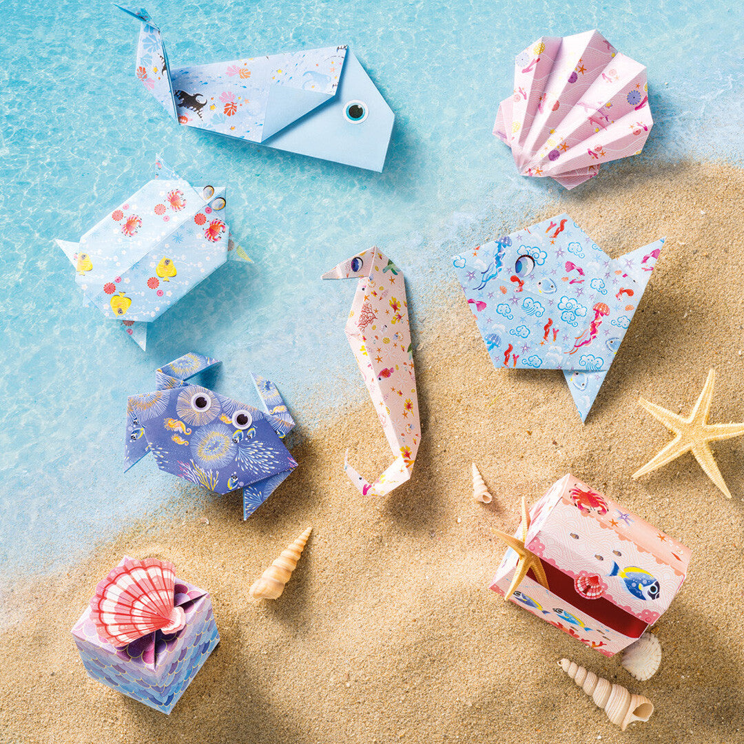 Diverse gevouwen origami zeedieren en doosjes uit de Janod Magic Ocean knutselset, liggend op een zandstrand met water en schelpen.