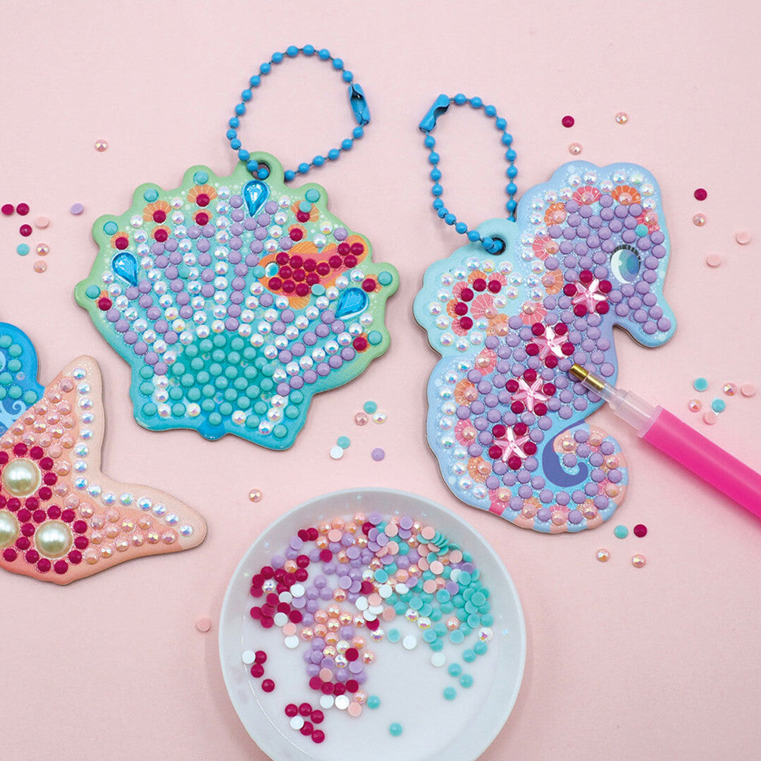 Twee afgewerkte Magic Ocean Diamond Painting sleutelhangers van een schelp en zeepaardje, met losse diamantjes en een aanbrengpen.
