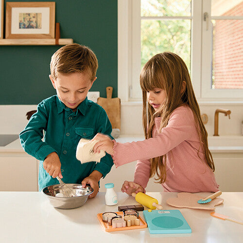 Twee kinderen spelen in een keuken met de Janod speelgoed bakset, waarbij de jongen klopt en het meisje 'bloem' toevoegt.