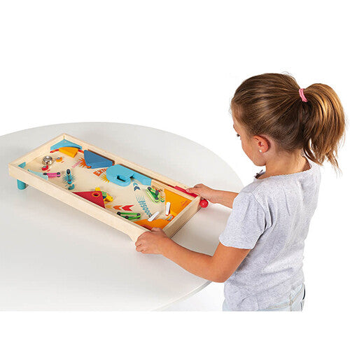 Een meisje staat aan een witte tafel en kijkt naar de Janod houten flipperkast.