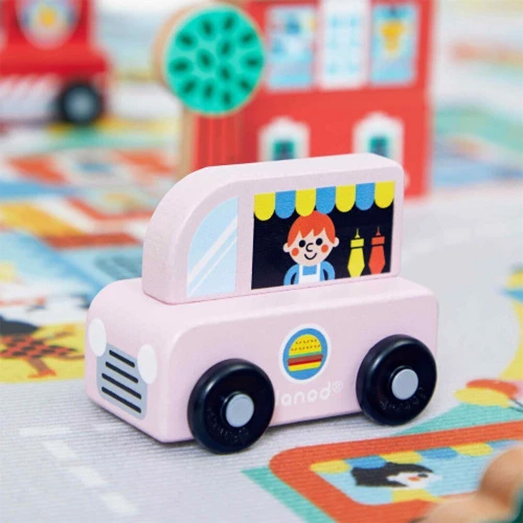 Janod roze houten speelgoed foodtruck op een speelmat met houten gebouwen op de achtergrond.