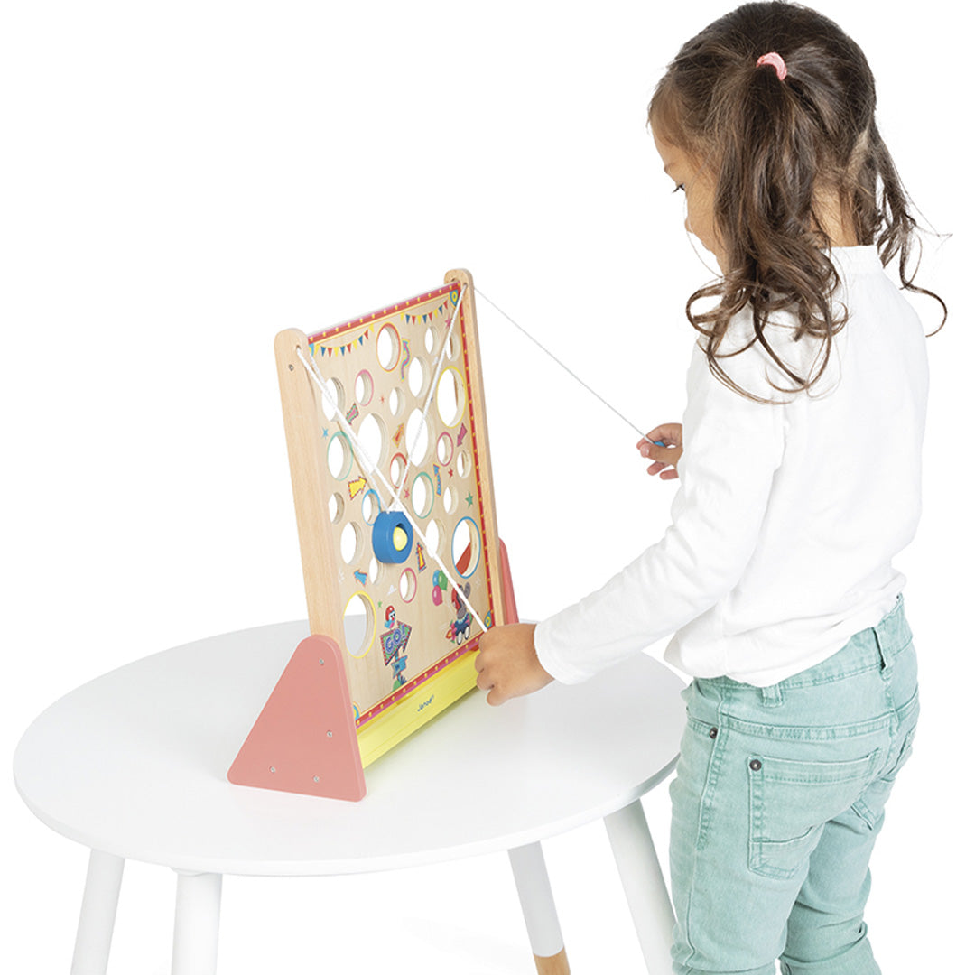 Een kind speelt met het Janod houten behendigheidsspel op een witte tafel, waarbij een gele bal met een touw door de gaten wordt geleid.