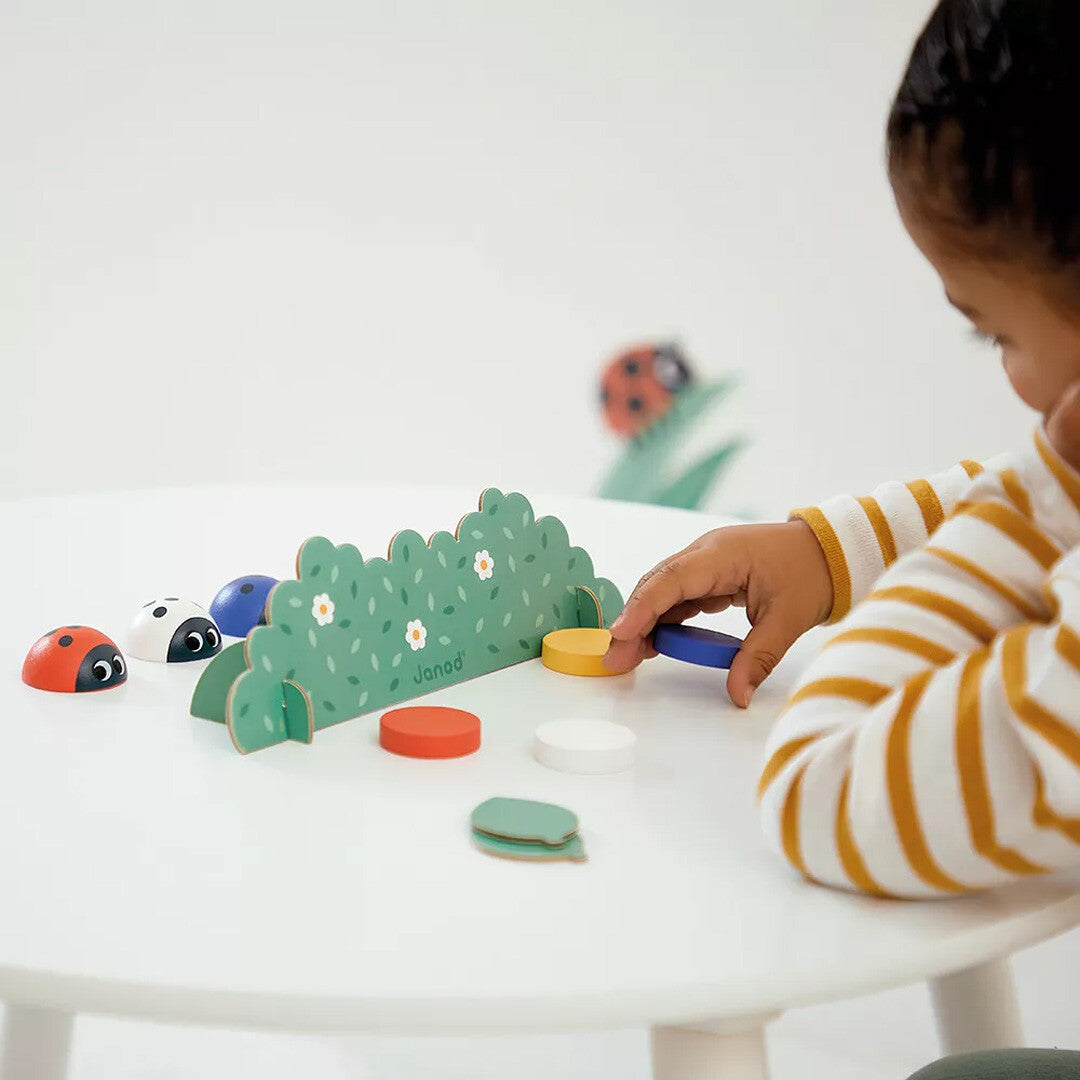 Een kind speelt met het Janod houten geheugenspel met lieveheersbeestjes en een groene struik.