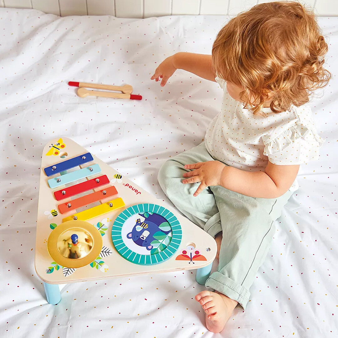 Een peuter met krullend haar zit op een wit bed en speelt met de Janod Gioia houten muziektafel.