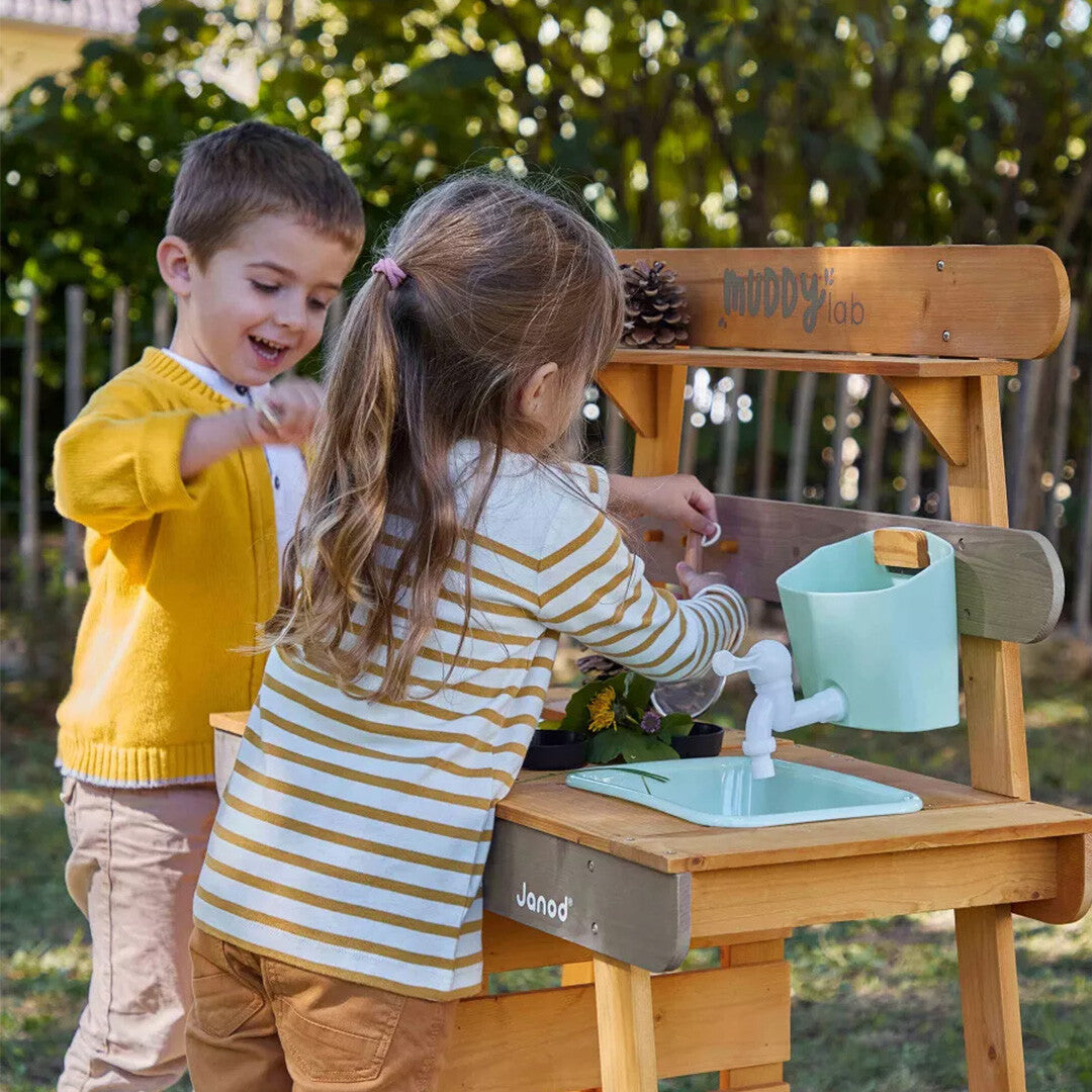 Twee kinderen spelen met de Janod houten modderkeuken in de tuin.