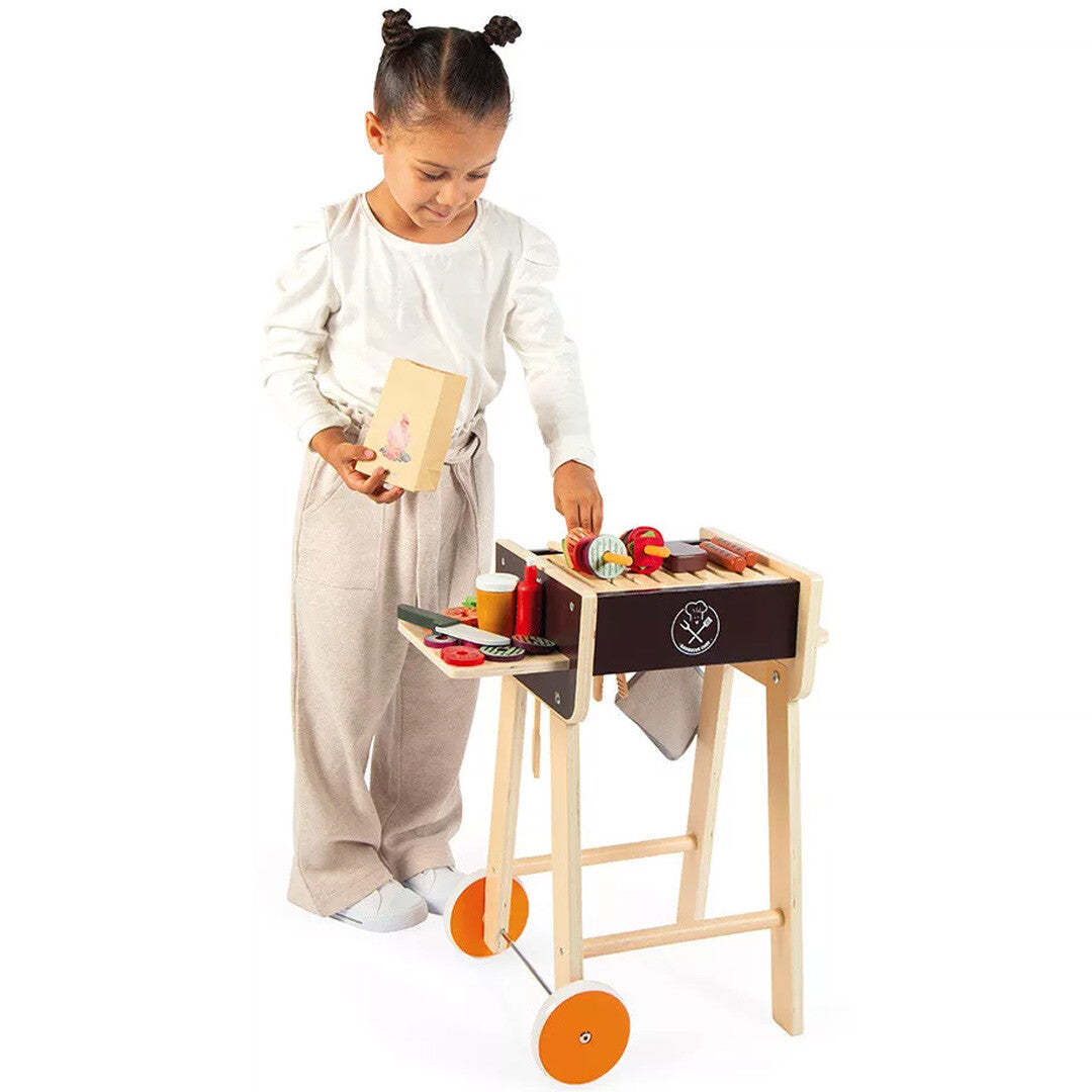 Een meisje speelt met de Janod houten kinderbarbecue en legt speelgoedeten op de grill.