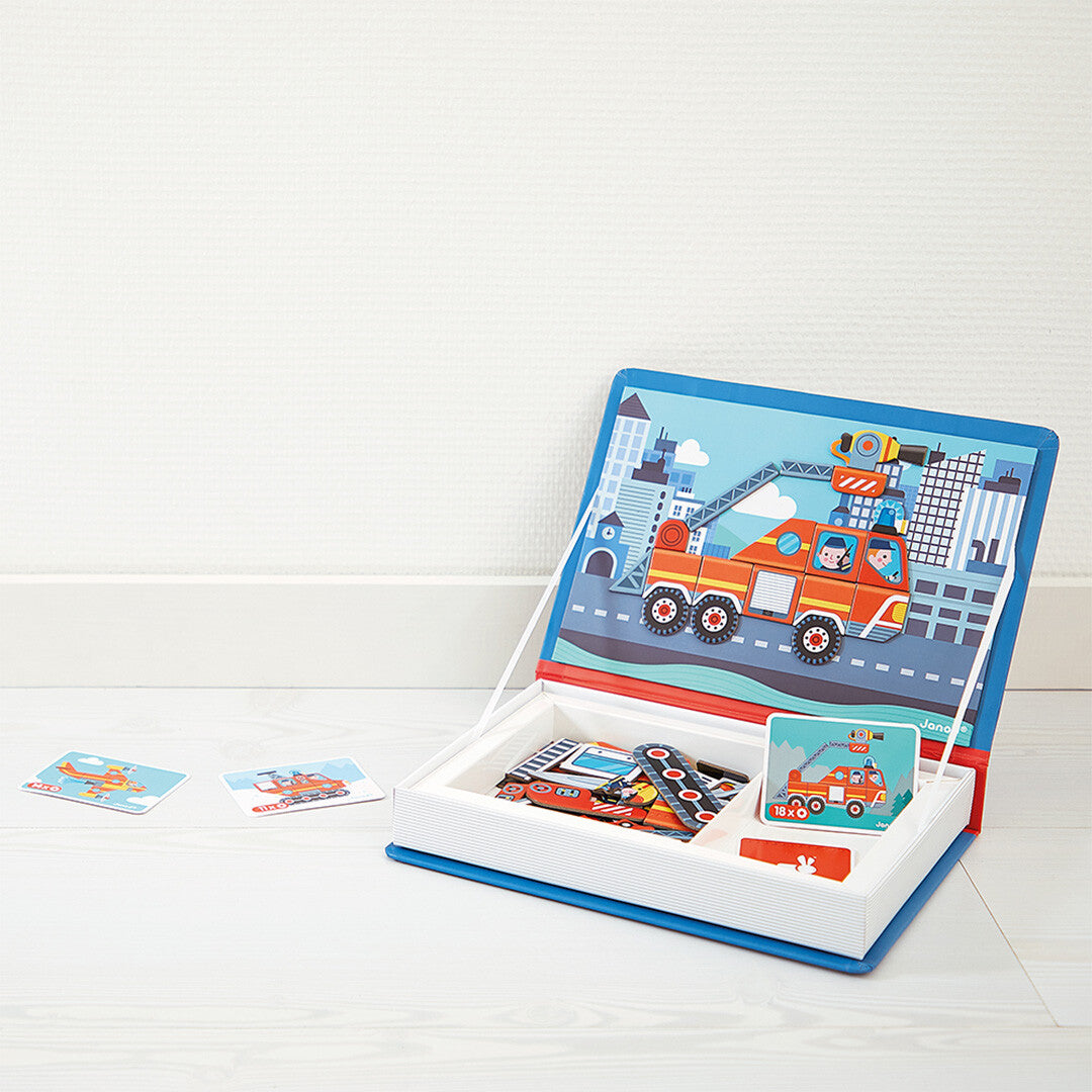 Het Janod magneetboek brandweer open op de vloer, met een brandweerwagen op de stadsachtergrond en twee voorbeeldkaarten ernaast.