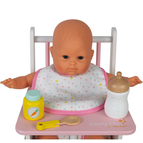 Een pop zit in een roze kinderstoel met een slab om, naast een flesje en potje uit de Janod poppen verzorgingsset.