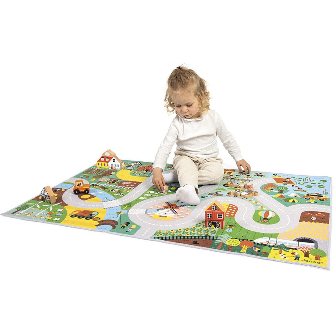 Een meisje zit op het Janod speelkleed 'Het Platteland' en speelt met houten speelgoed.