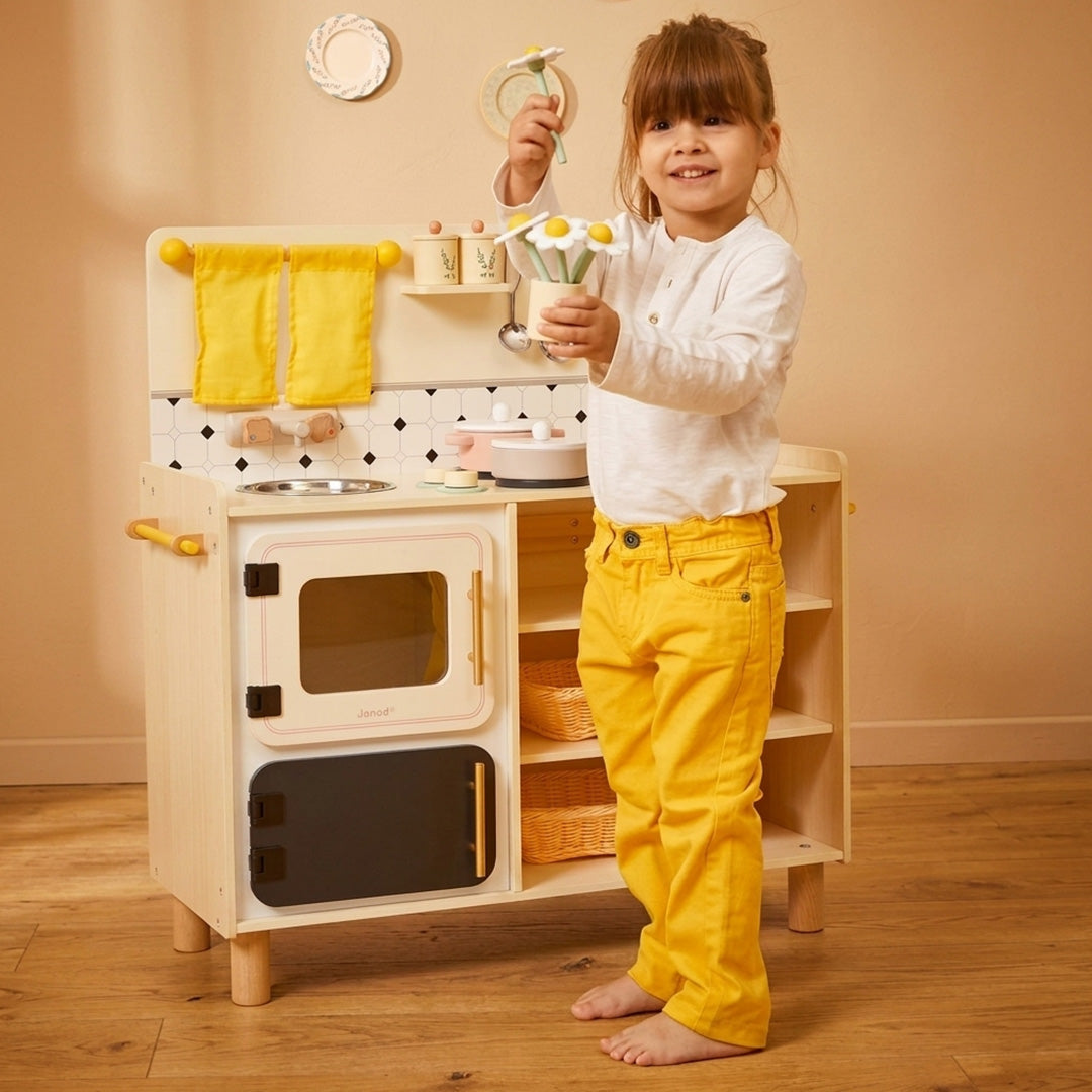 Een kind staat naast de Janod kinderkeuken en houdt speelgoedbloemen vast.