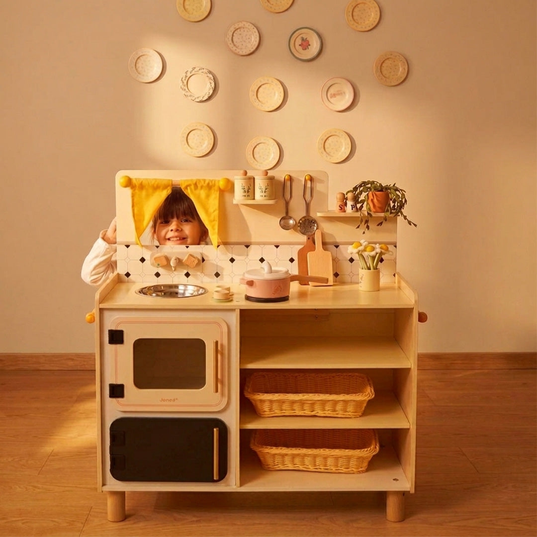 Een kind kijkt lachend over de Janod kinderkeuken heen.