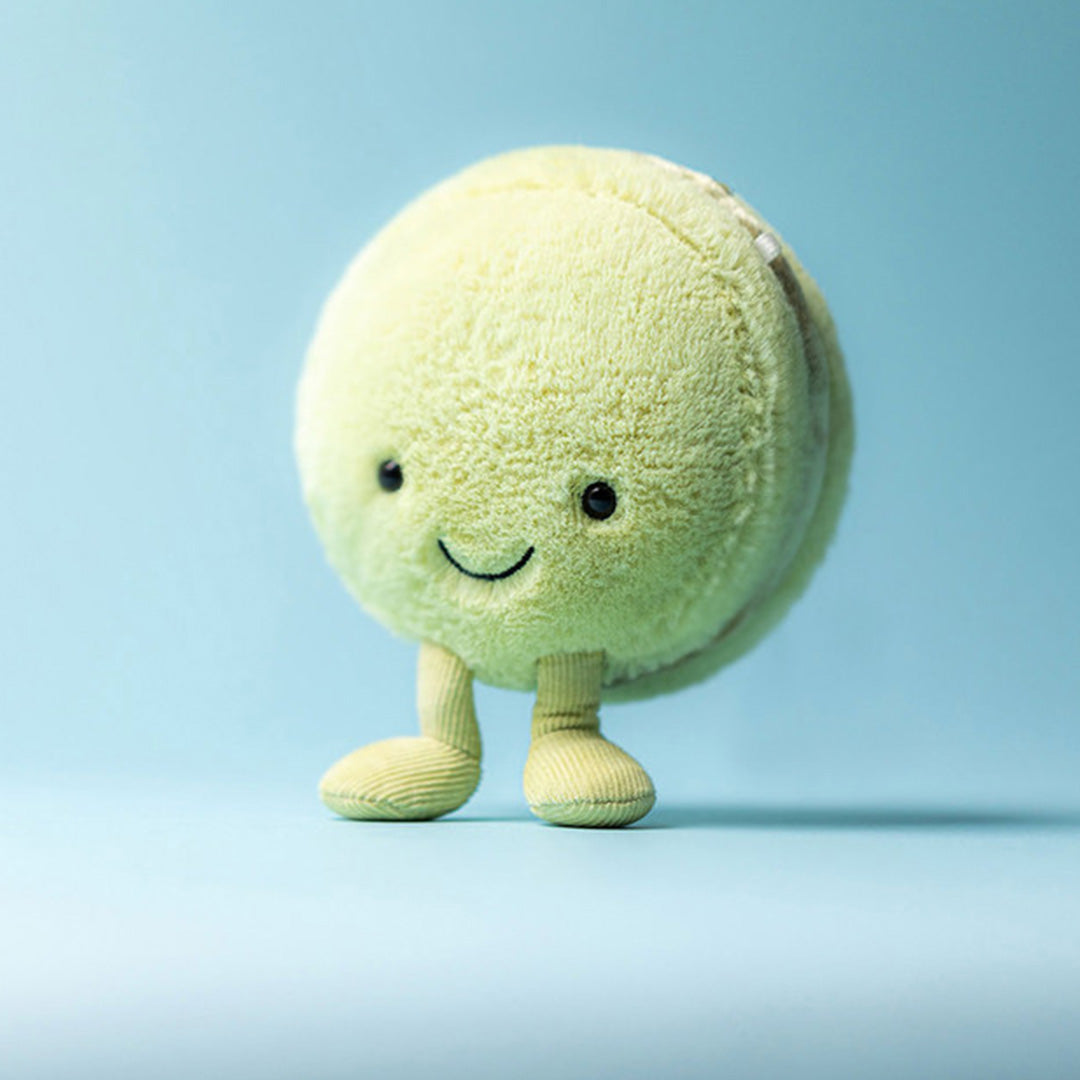 Jellycat pluchen pistache macaron knuffel met lachend gezicht en kleine voetjes, staand op een blauwe achtergrond.