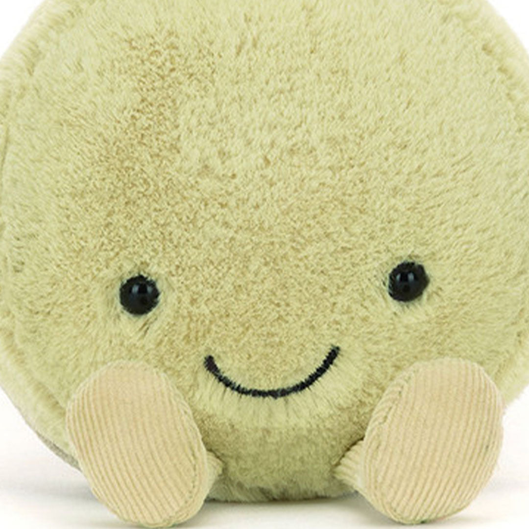 Close-up van het lachende gezicht van de Jellycat pluchen pistache macaron knuffel met zwarte ogen en kleine handjes.