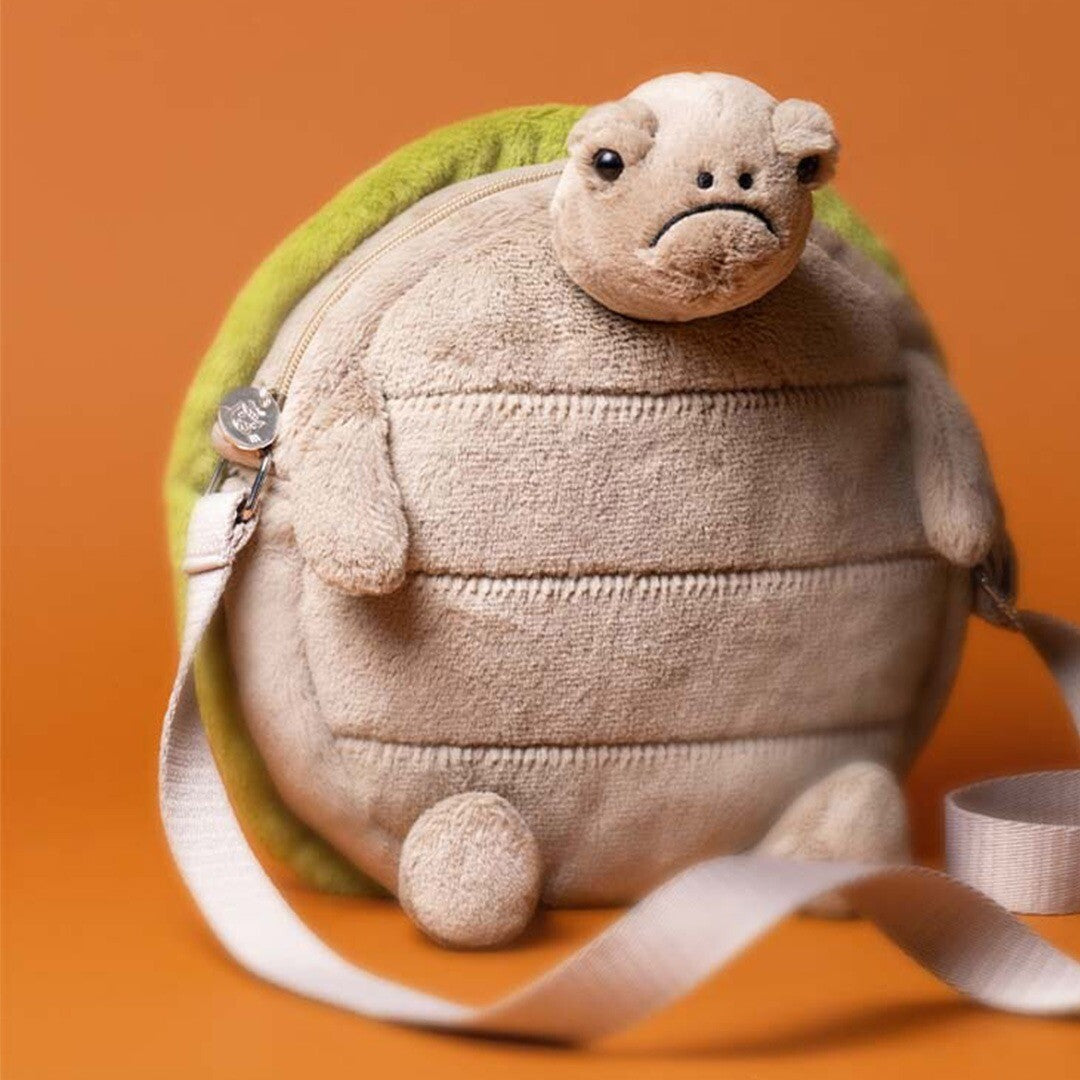 Jellycat kindertas in de vorm van een schildpad met beige lijf en groene schild, schuin van voren gezien tegen een oranje achtergrond.