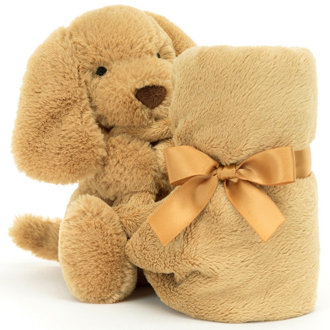 Jellycat knuffeldoek hond toffee, een zittende pluchen hond die een opgerold bruin dekentje met gouden lint vasthoudt.