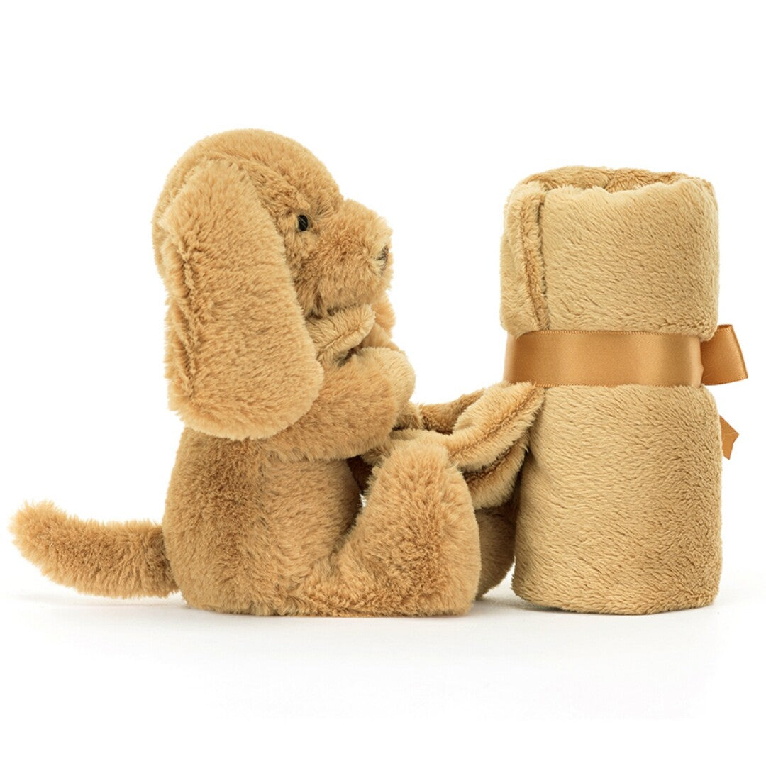Zijaanzicht van de Jellycat knuffeldoek hond toffee, een zittende pluchen hond naast een opgerold bruin dekentje.