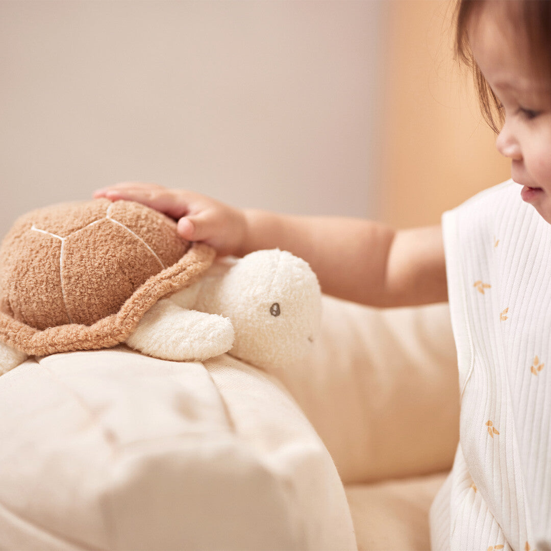 Een kinderhand aait de Jollein activiteitenknuffel schildpad die op een lichte ondergrond ligt.