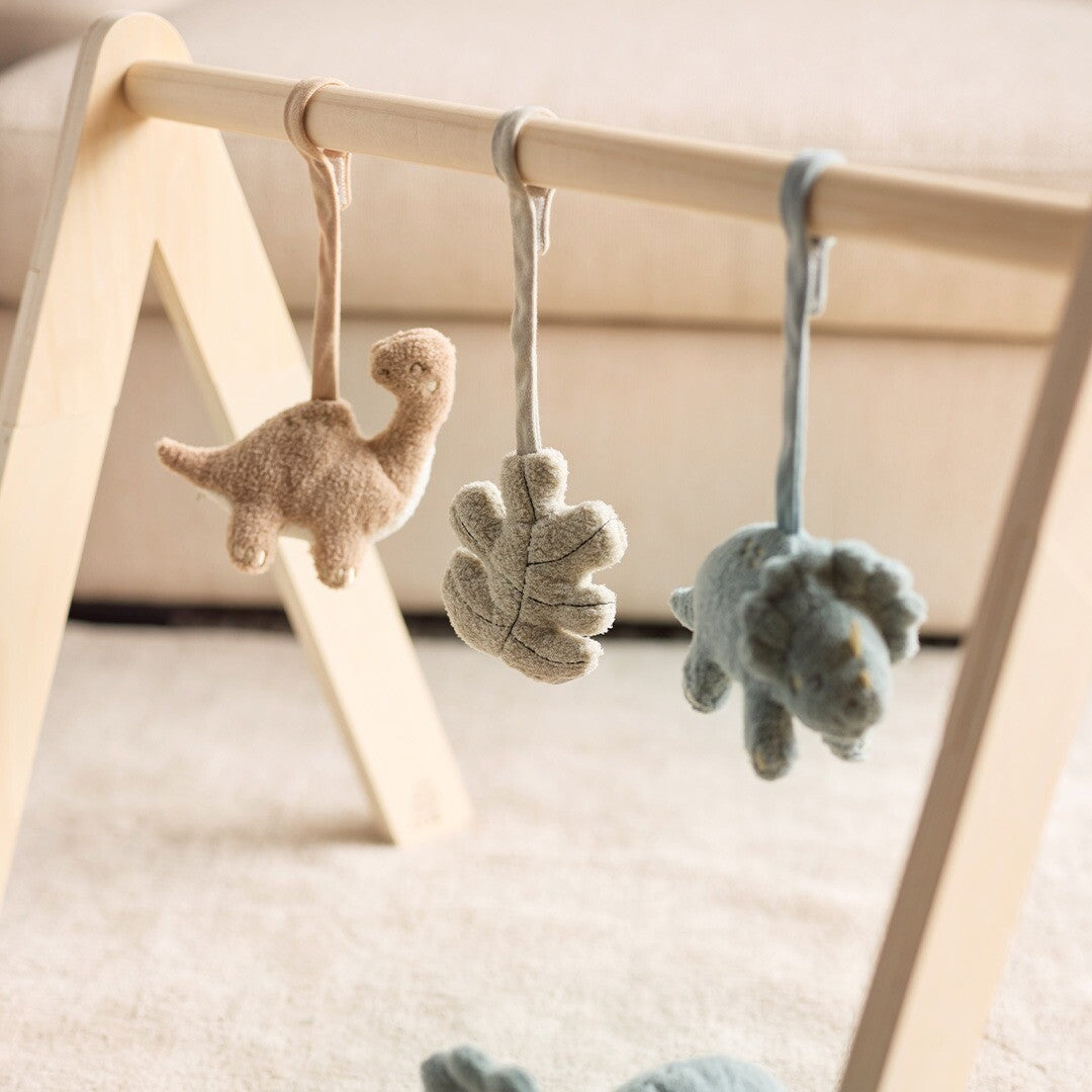Jollein babygymspeeltjes met een beige dinosaurus, een beige blad en een blauwe dinosaurus hangend aan een houten babygym op een lichte vloer.