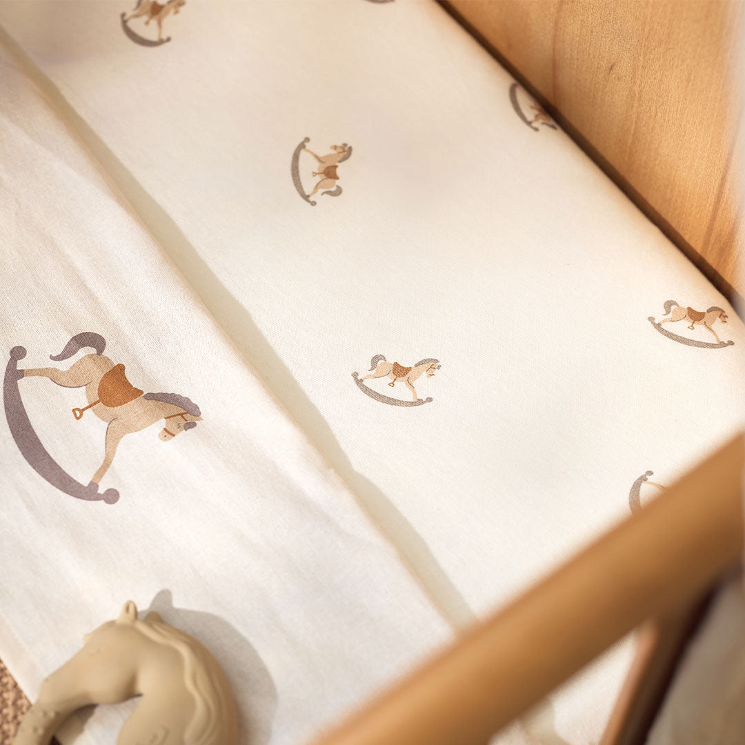 Jollein babyhoeslaken met beige wiebelpaardjespatroon in een ledikant, met een bijtring op de voorgrond.