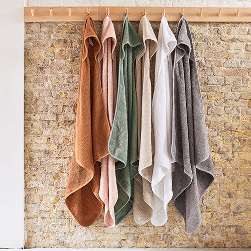 Jollein badcapes in diverse kleuren hangen aan een houten rek tegen een bakstenen muur.