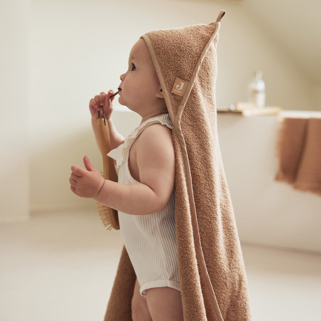 Baby met een Jollein badcape van badstof in biscuitkleur op het hoofd, staand met een borstel.